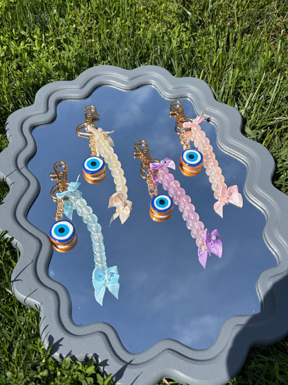 Four beaded keychains with evil eye charms are displayed on a decorative gray tray outdoors on green grass. The keychains come in light blue, cream, pink, and lavender, each with a gold-tone clasp and a small ribbon bow at the bottom.