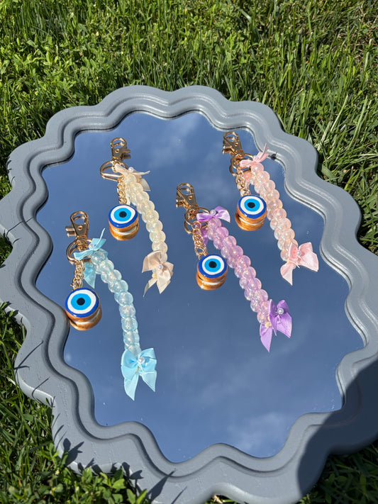 Four beaded keychains with evil eye charms are displayed on a decorative gray tray outdoors on green grass. The keychains come in light blue, cream, pink, and lavender, each with a gold-tone clasp and a small ribbon bow at the bottom.